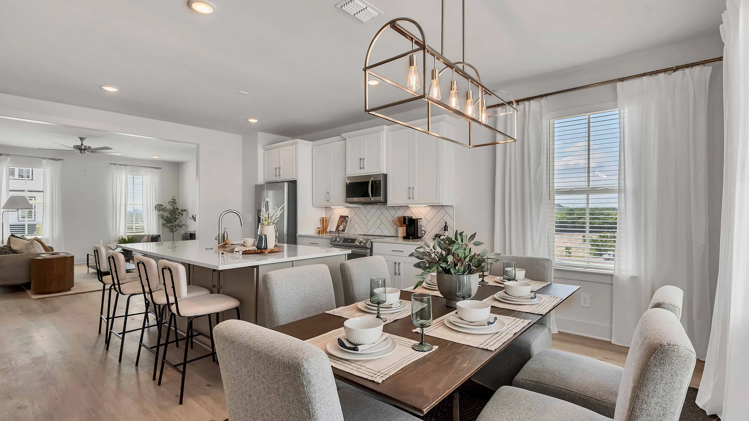 Modern open-plan kitchen and dining area with elegant decor, featuring a spacious island with bar seating, chic lighting, and a large window with natural light.