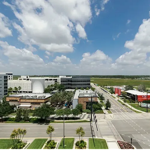Vibrant urban view showcasing modern buildings, lush greenery, and a wide street under a bright blue sky with fluffy clouds.