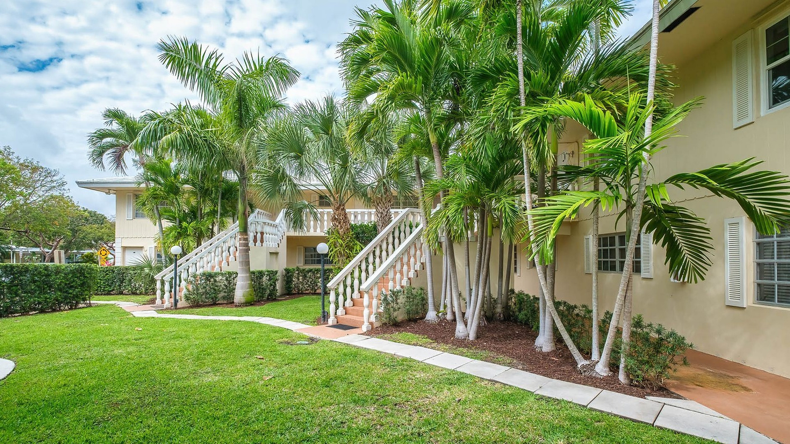 Beautiful tropical apartment exterior with lush palm trees and elegant stairs, surrounded by manicured greenery.