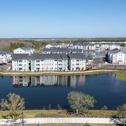 Aerial view of modern apartment complex nestled by a serene lake, surrounded by lush greenery and suburban charm.