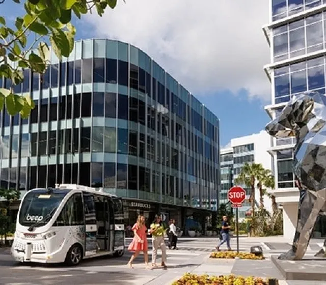 Modern urban scene featuring sleek glass office buildings, automated shuttle bus, and striking metallic dog sculpture under a vibrant sky.