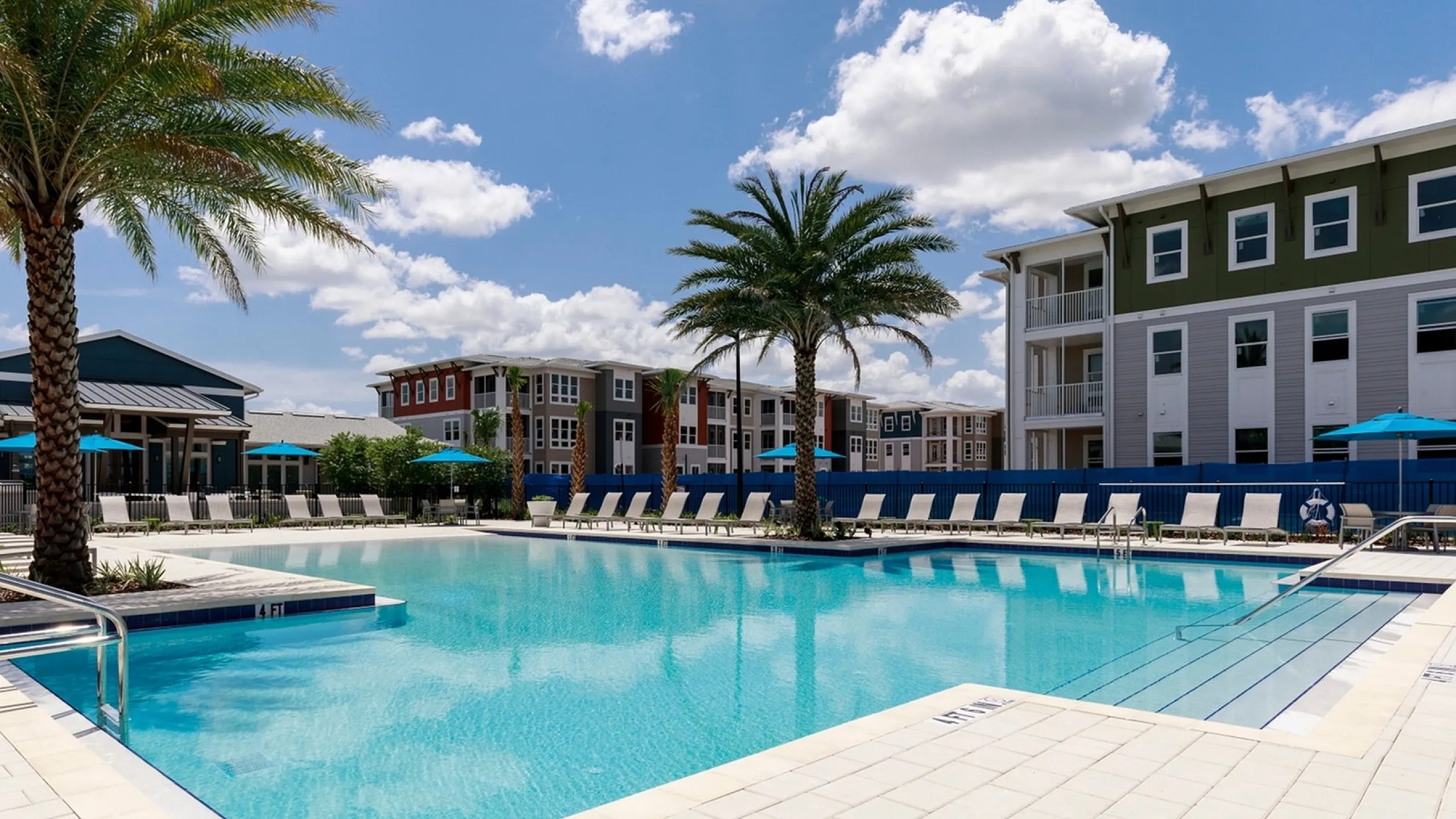 Luxurious apartment complex featuring a sparkling outdoor pool surrounded by lush palm trees and modern buildings, ideal for relaxation under clear blue skies.