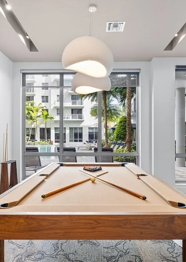 Sleek and modern apartment lobby featuring a stylish pool table, elegant pendant lighting, and large windows with views of a lush courtyard.