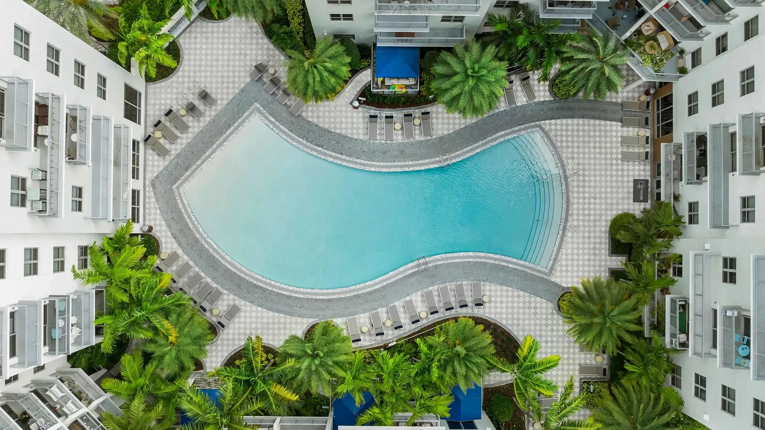 Bird's-eye view of a luxurious residential complex featuring a stunning, curved swimming pool surrounded by elegant palm trees and sun loungers, offering a relaxing oasis amidst modern apartment buildings.