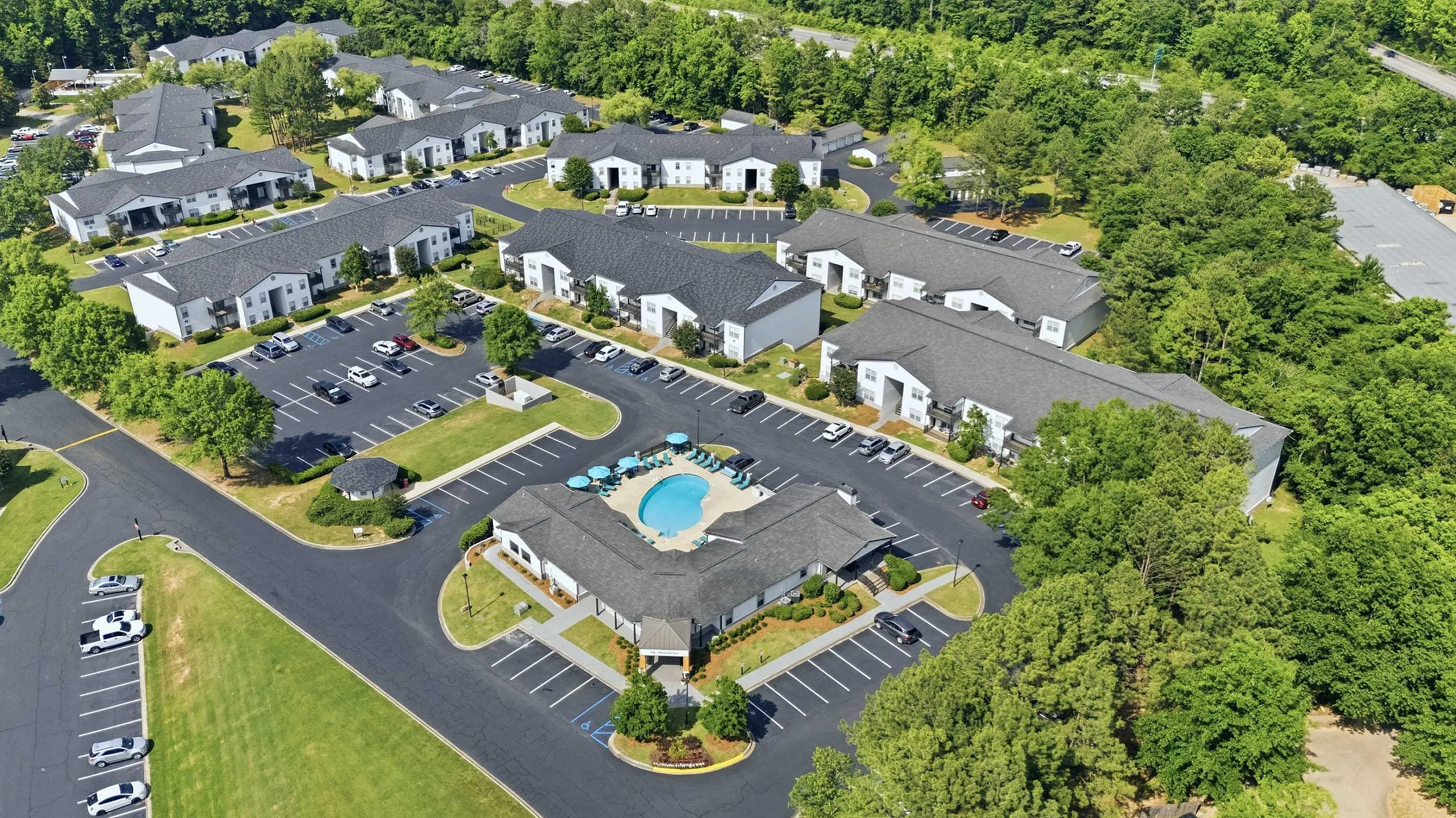 Aerial view of a modern apartment complex with a central pool, abundant greenery, and ample parking spaces for a comfortable lifestyle.