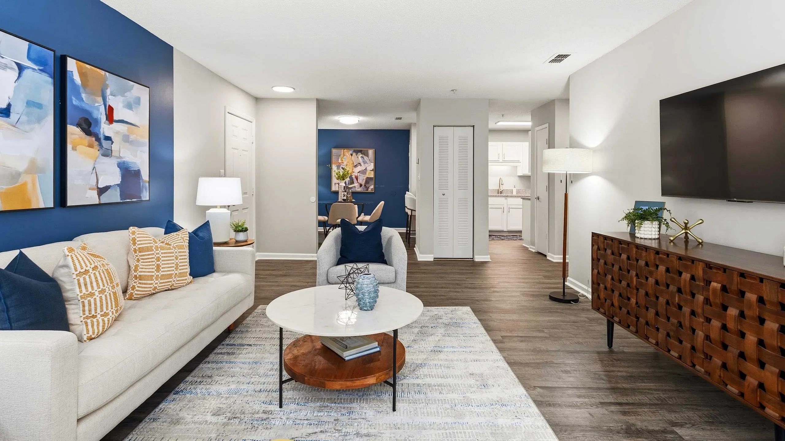 Modern living room with elegant decor, featuring a cozy sofa, artistic wall paintings, and a view into a stylish dining area and kitchen.