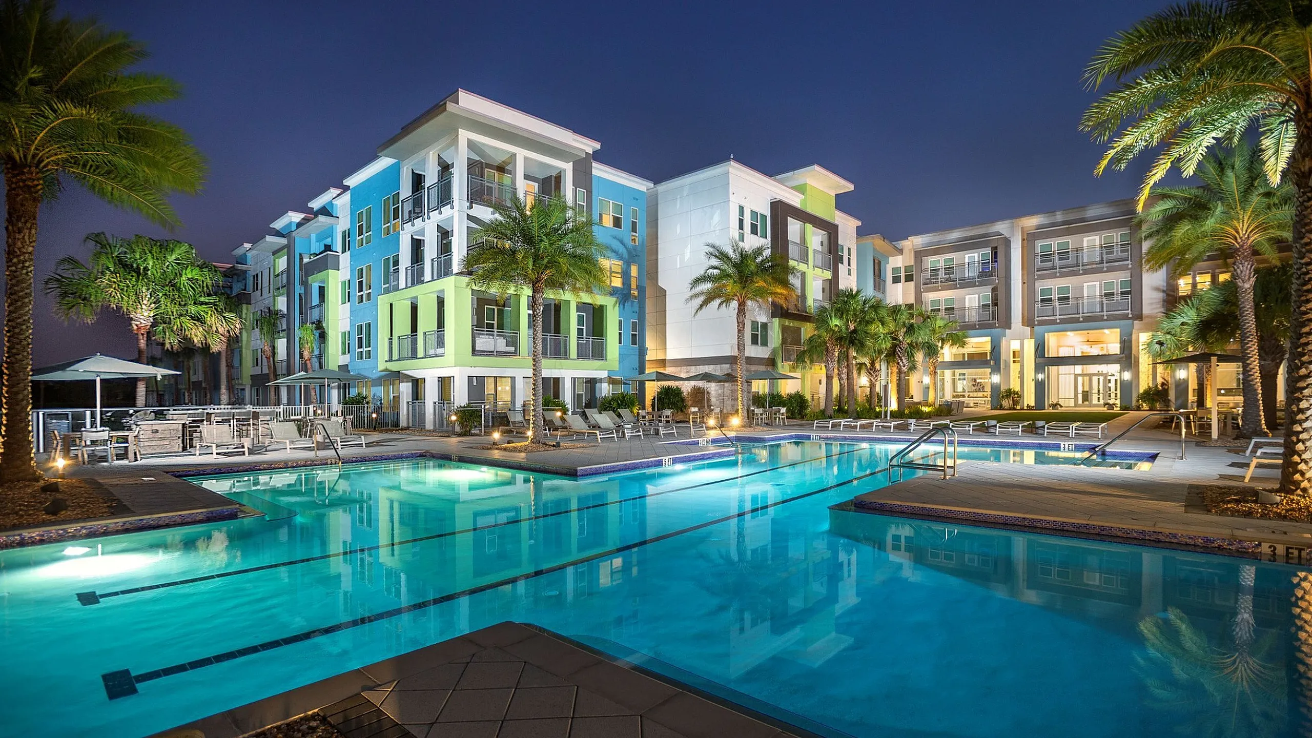 Luxurious apartment complex with vibrant colors, surrounded by palm trees, featuring a stunning illuminated pool area at night.