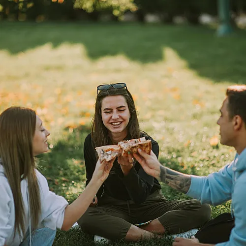 Joyful friends enjoying a sunny picnic in the park, sharing slices of delicious pizza on a vibrant, grassy lawn.