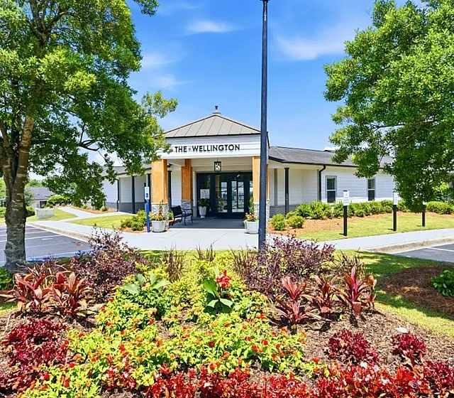 Charming entrance of The Wellington, surrounded by vibrant landscaped gardens and mature trees under a clear blue sky.