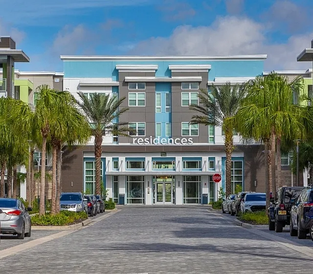 Modern residential building entrance surrounded by lush palm trees and parked cars, showcasing vibrant architecture under a clear blue sky.