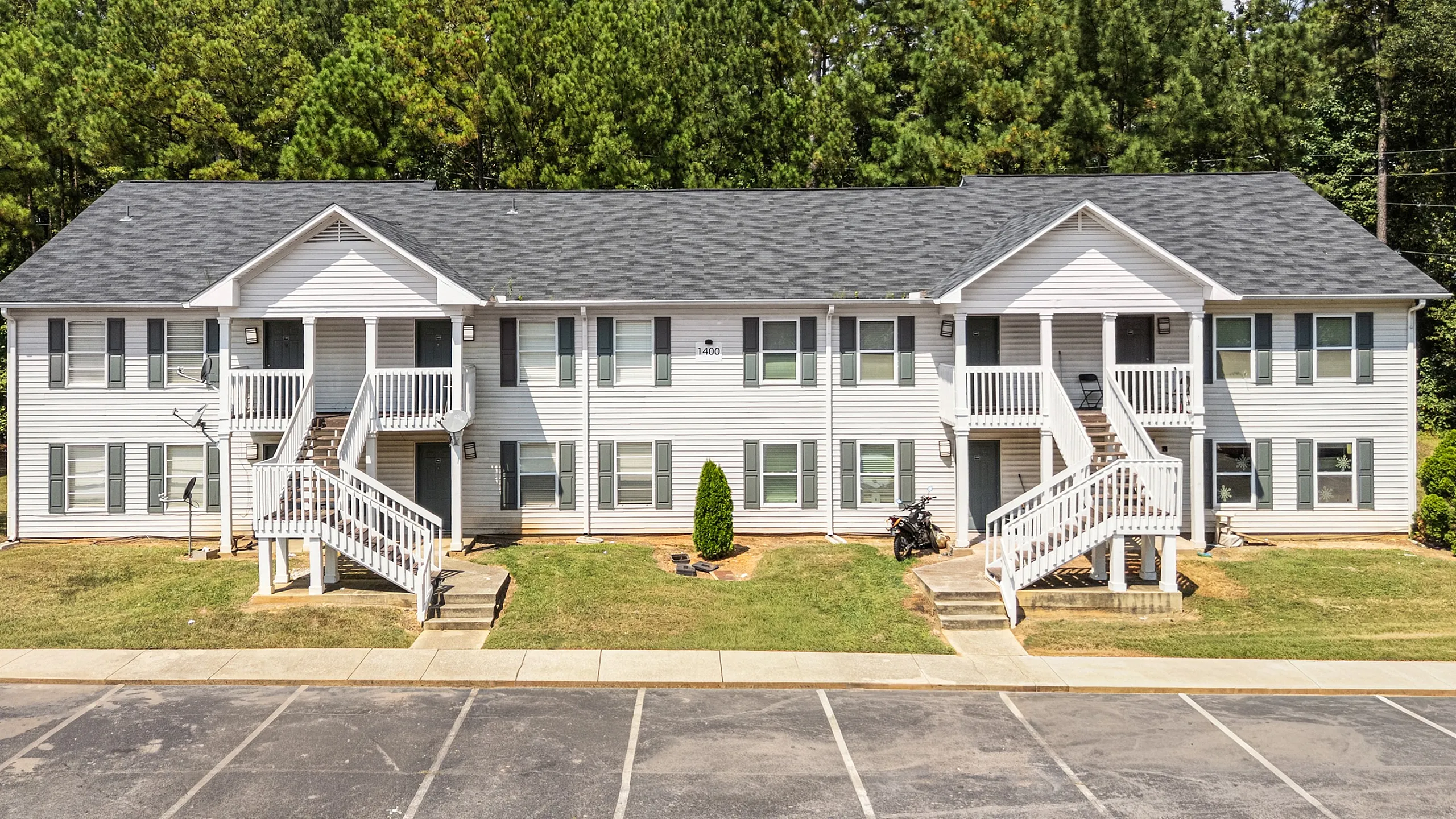 Charming two-story apartment building with white siding and inviting front porches set against a lush green backdrop.