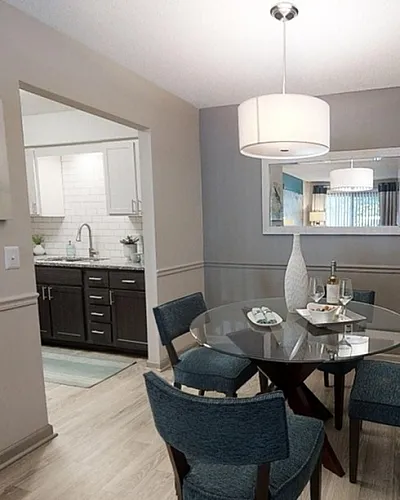 Elegant dining room with a modern glass table, stylish chairs, and contemporary artwork, leading into a sleek kitchen with dark wood cabinetry and bright lighting.