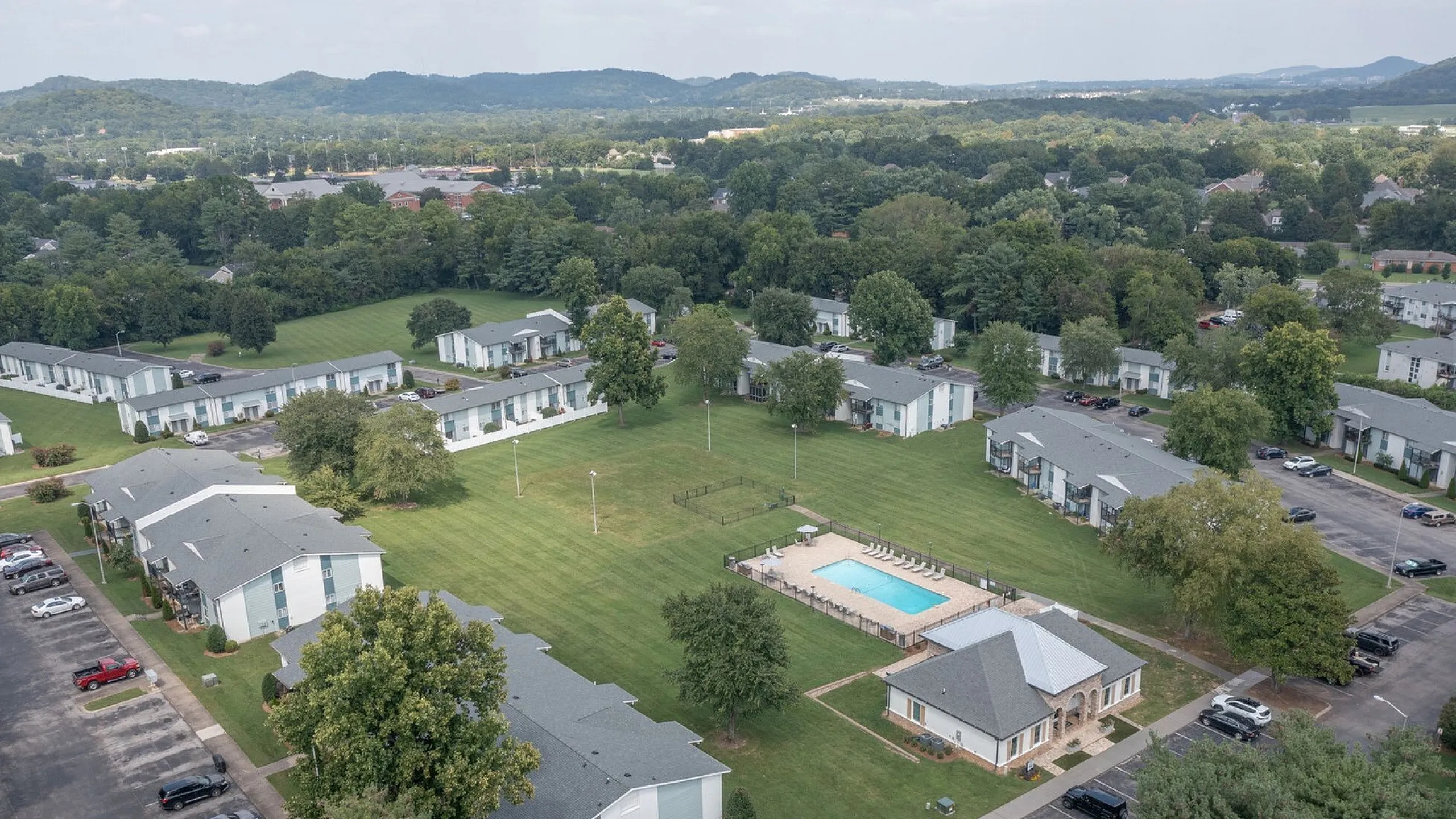 Aerial view of a charming apartment complex featuring lush green lawns, modern buildings, and a central sparkling pool surrounded by scenic landscape.