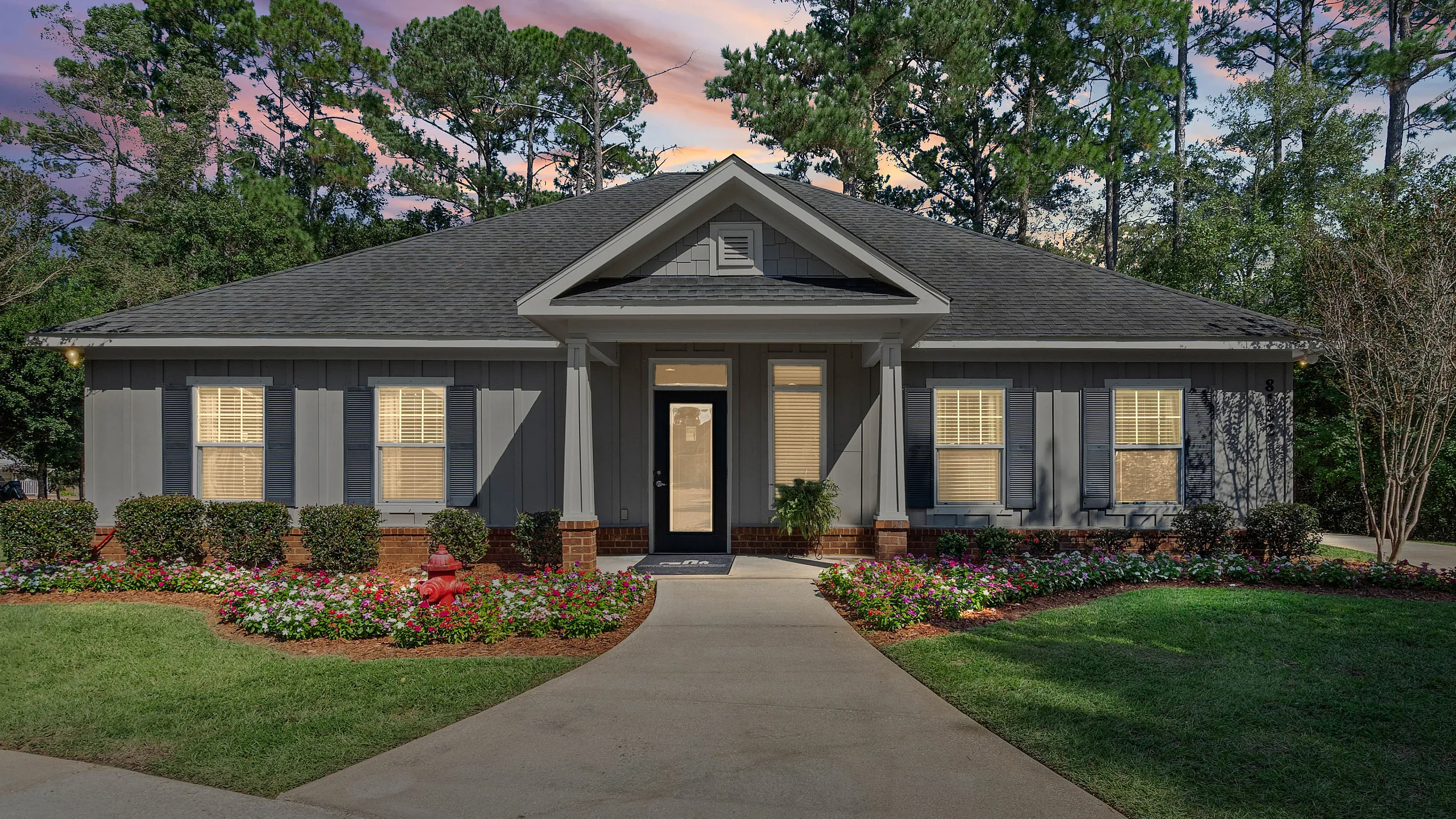 Charming single-story home with elegant landscaping and a welcoming front porch, set against a serene backdrop of lush trees and a vibrant sunset sky.