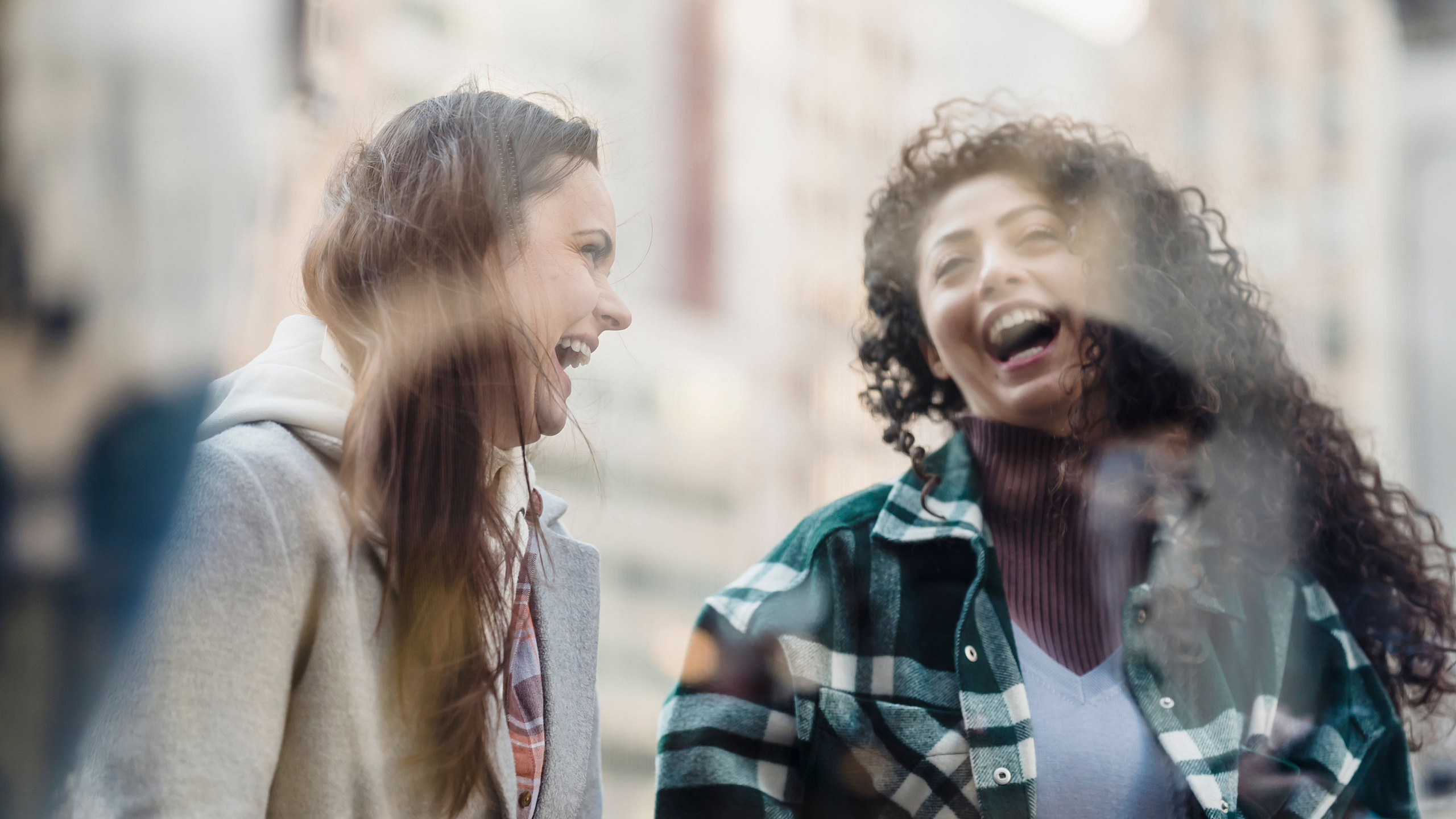 Joyful encounter of two friends laughing together, captured through a city cafe's window reflection.