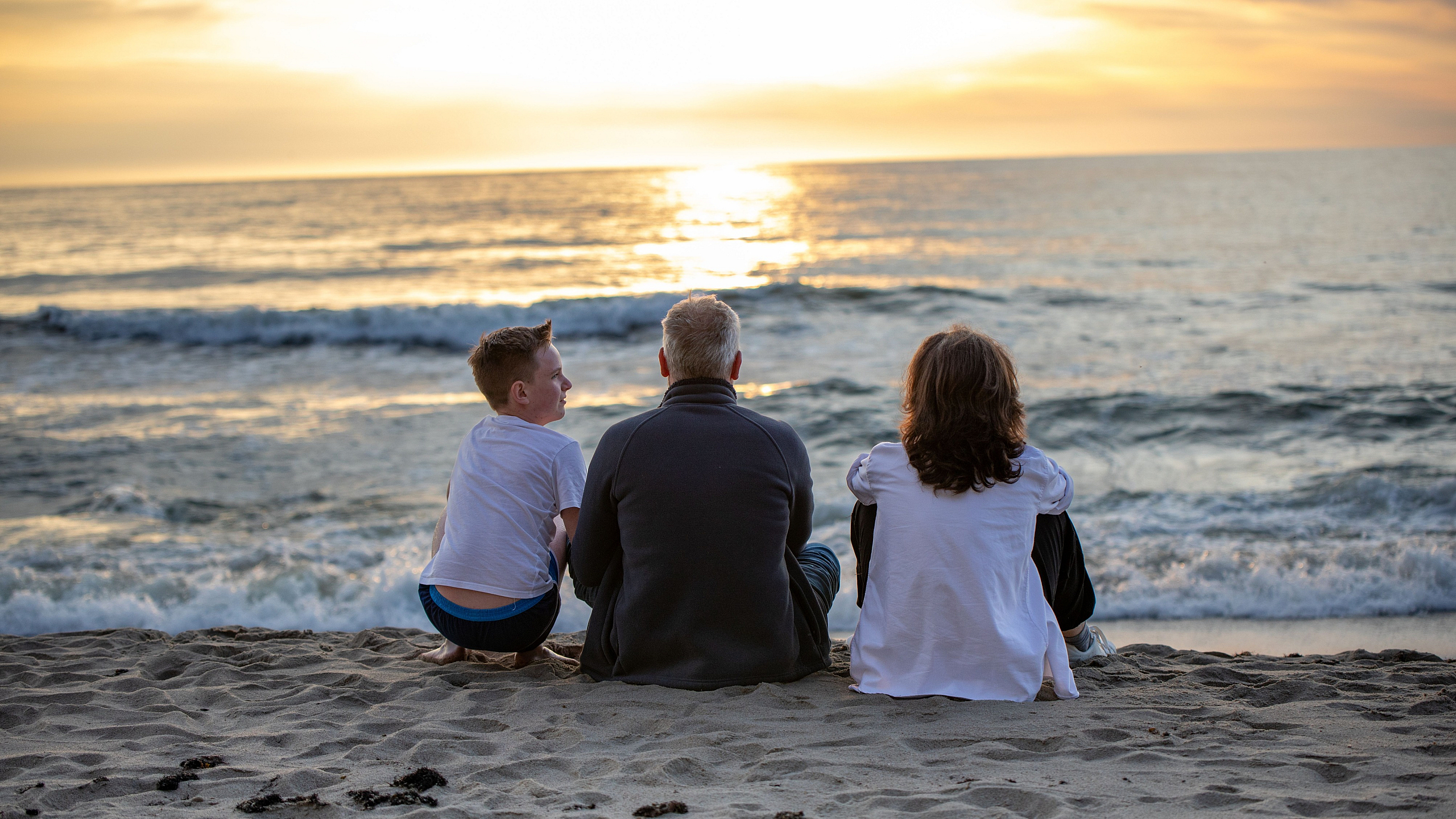 Serene beach sunset with three people sitting on the sand, enjoying the gentle waves and golden horizon.