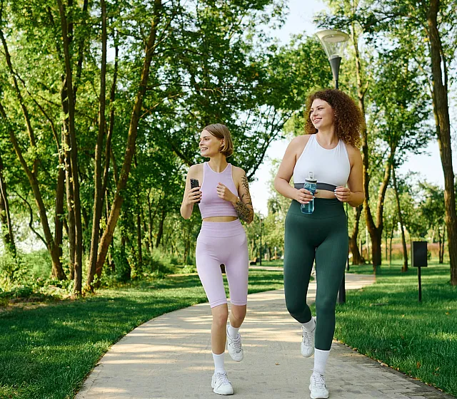 Two women jogging on a lush, tree-lined path, enjoying an outdoor workout in fresh air.