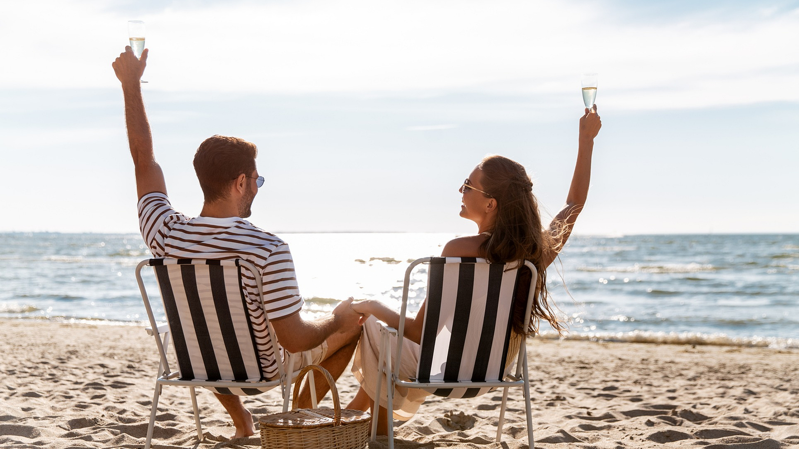 Couple in striped chairs enjoying a sunny day on the beach, toasting with glasses of champagne, with a serene ocean backdrop.