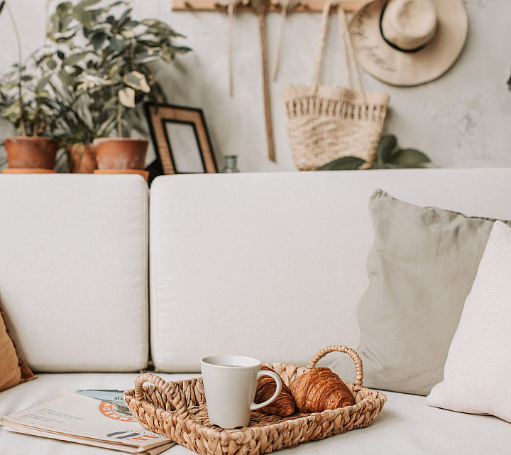 Inviting living area with cozy white sofa adorned with pastel cushions, complemented by lush green plants and stylish decor. A rustic basket holds a warm cup of coffee and croissants on the sofa.