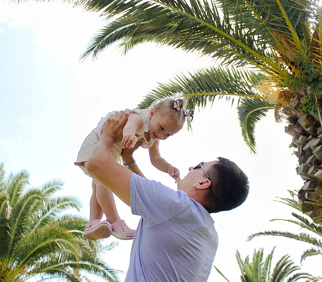 Joyful outdoor moment of a man playfully lifting a child, surrounded by lush palm trees under a bright blue sky.