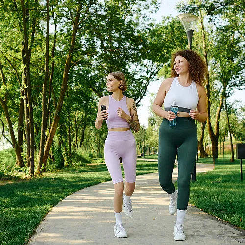 Two women jogging joyfully on a tree-lined path in a vibrant green park, enjoying a healthy lifestyle outdoors.