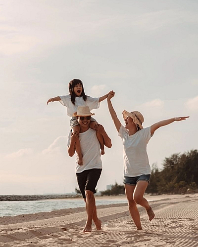 Joyful family enjoying a sunny day at the beach, with a child on shoulders and parents holding hands, along a scenic shoreline.