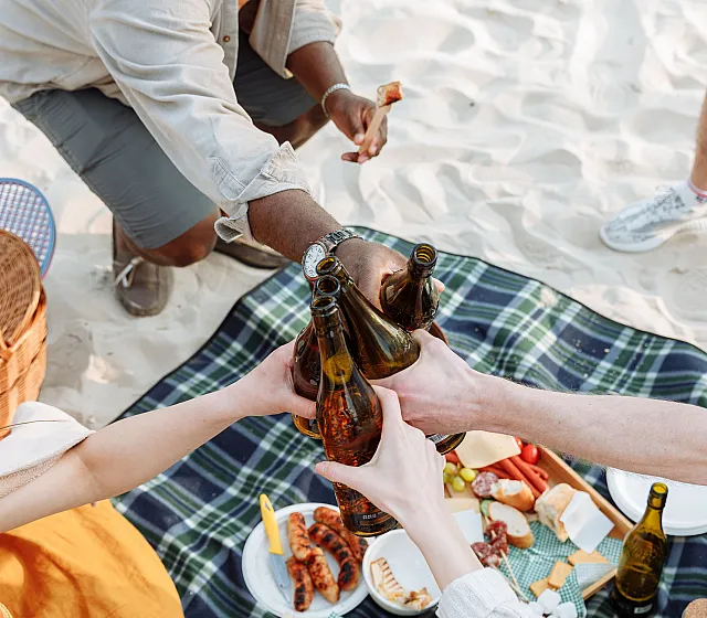 Joyful beach picnic scene with friends toasting with drinks over a plaid blanket, surrounded by a delicious spread of snacks and a wicker basket.