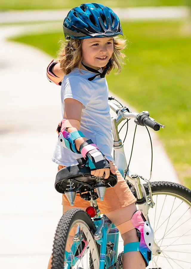 Joyful child riding a bicycle on a sunny path, wearing a helmet and colorful safety gear, surrounded by lush green grass.