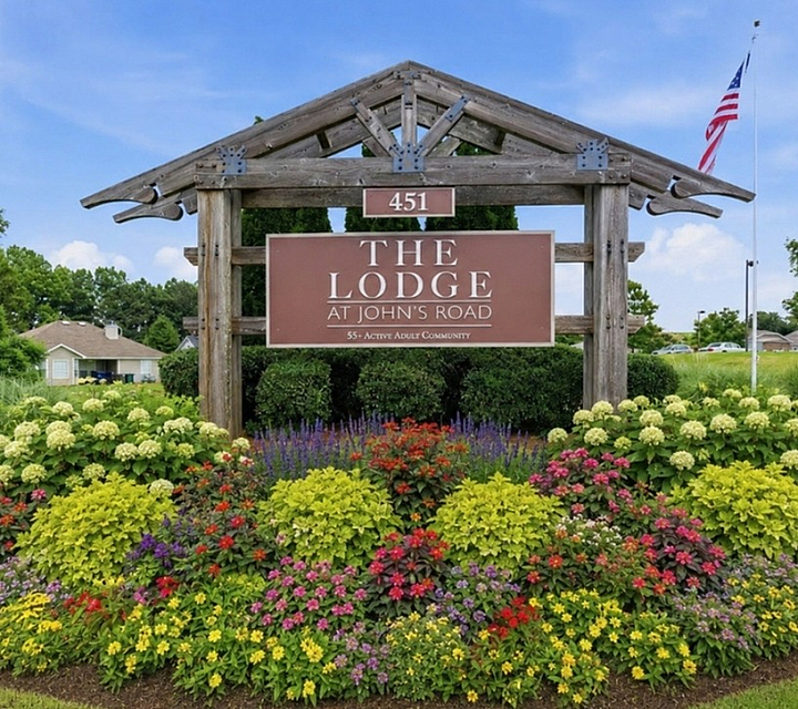 Welcome to The Lodge at John's Road, an inviting entrance surrounded by vibrant, colorful flower gardens and lush greenery, perfect for an active adult community.