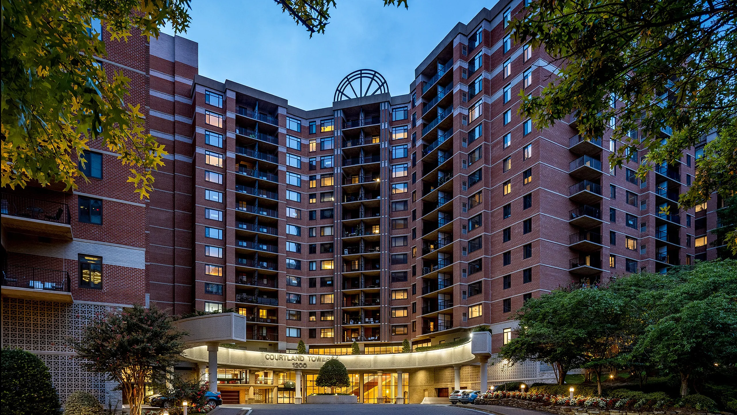 Elegant high-rise apartment building with numerous balconies, surrounded by lush greenery, illuminated beautifully at dusk, offering modern urban living.