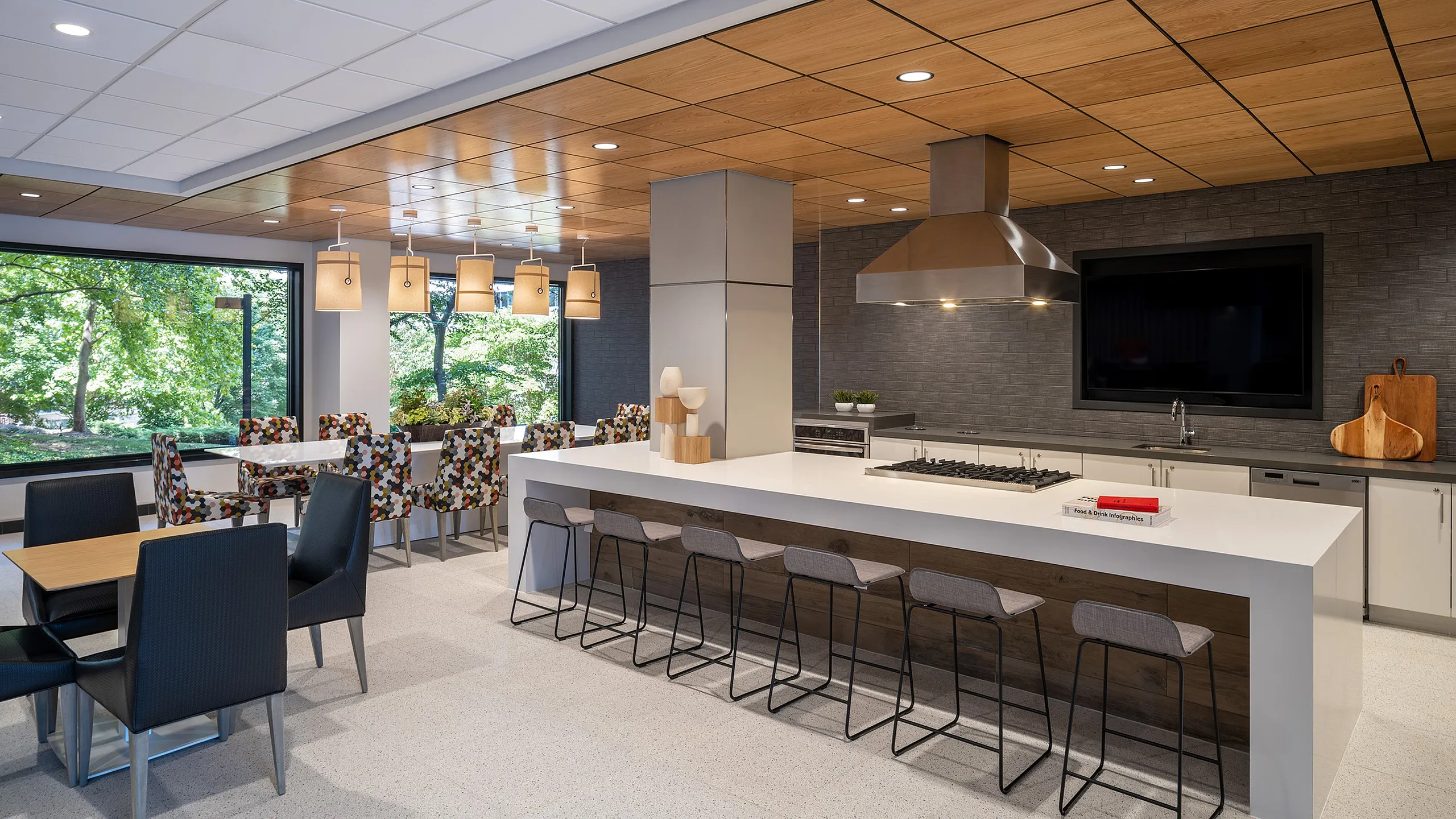 Modern communal kitchen space featuring sleek countertops, stylish bar stools, and vibrant dining chairs under warm pendant lighting, with large windows offering a lush green view.