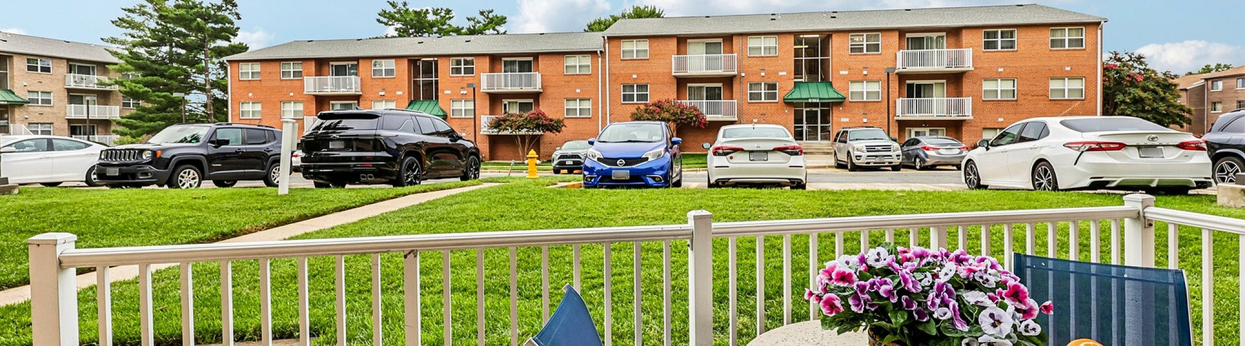 Charming apartment balcony with patio furniture and vibrant flowers overlooking a well-maintained lawn and modern apartment buildings.