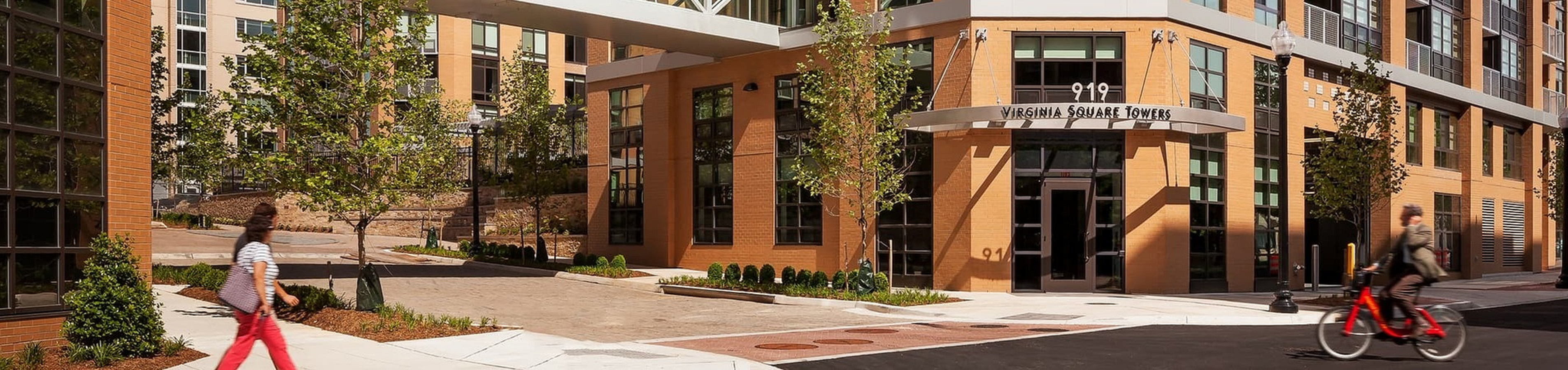 Modern urban apartment complex with distinctive brick design and glass walkway, surrounded by trees and vibrant city life.