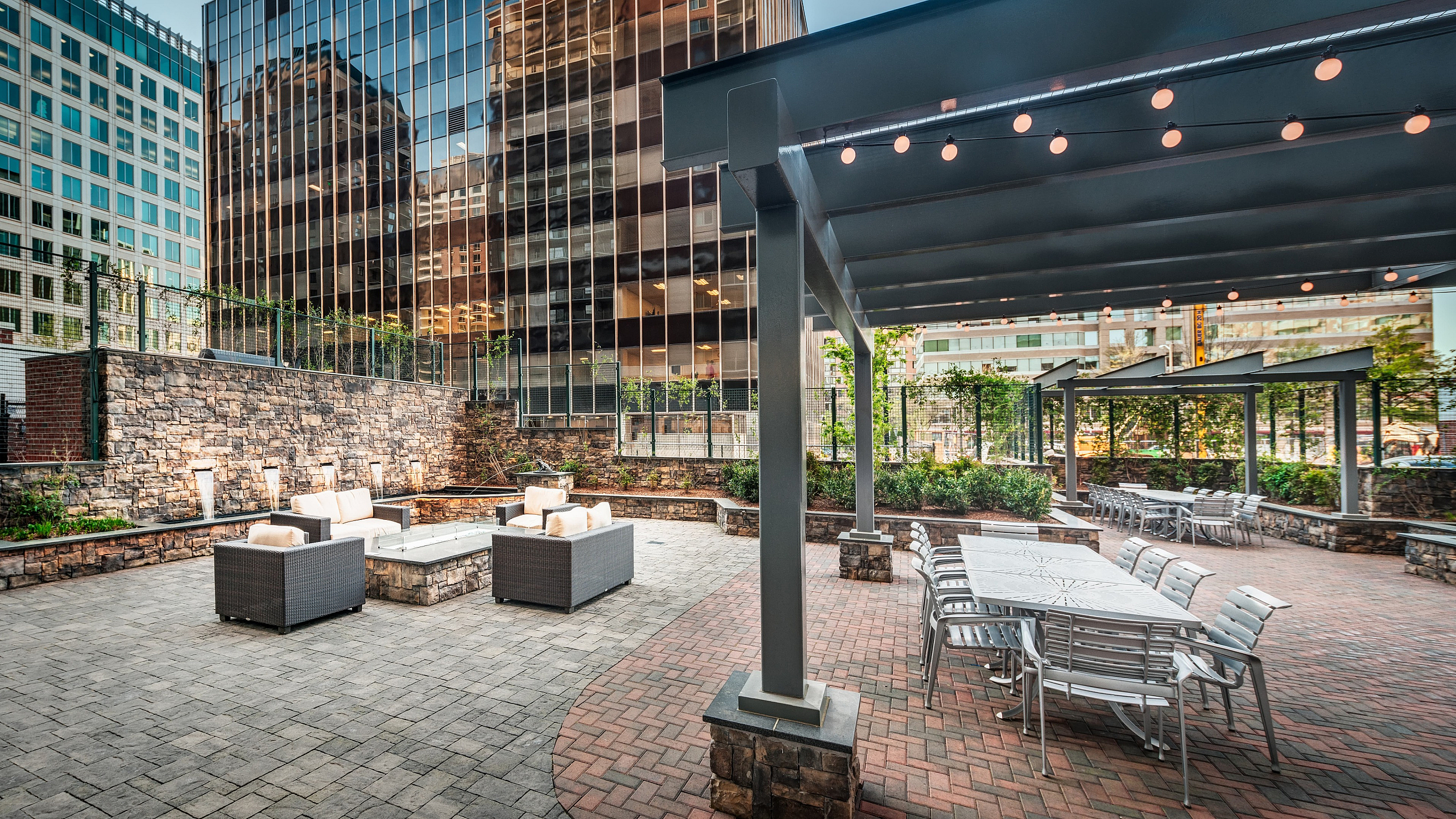 Urban oasis with elegant outdoor seating, string lights, and lush greenery surrounded by modern high-rise buildings.