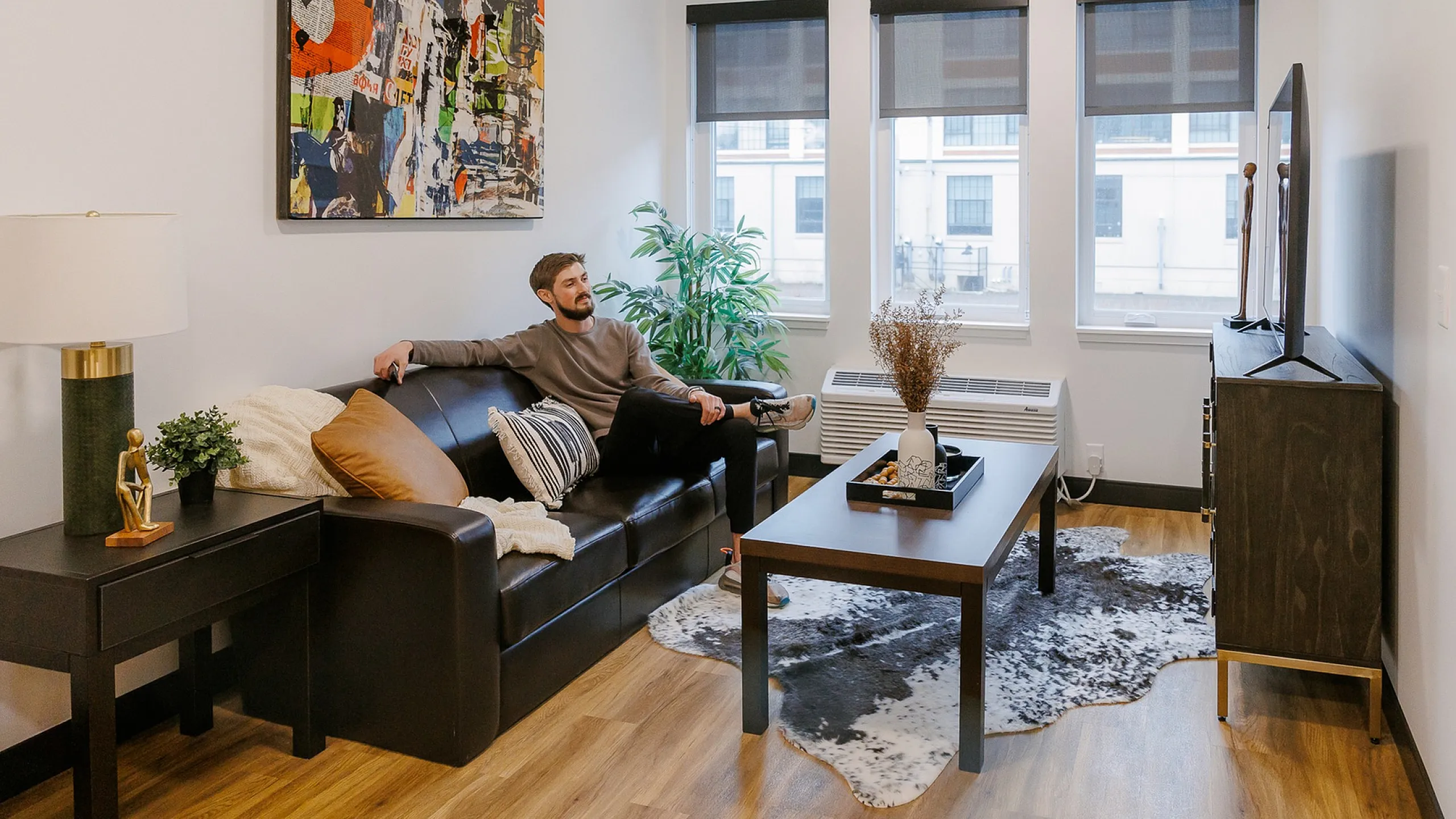 Cozy and inviting living room with stylish decor, featuring a comfortable sofa, artistic wall painting, and large windows bringing in natural light.