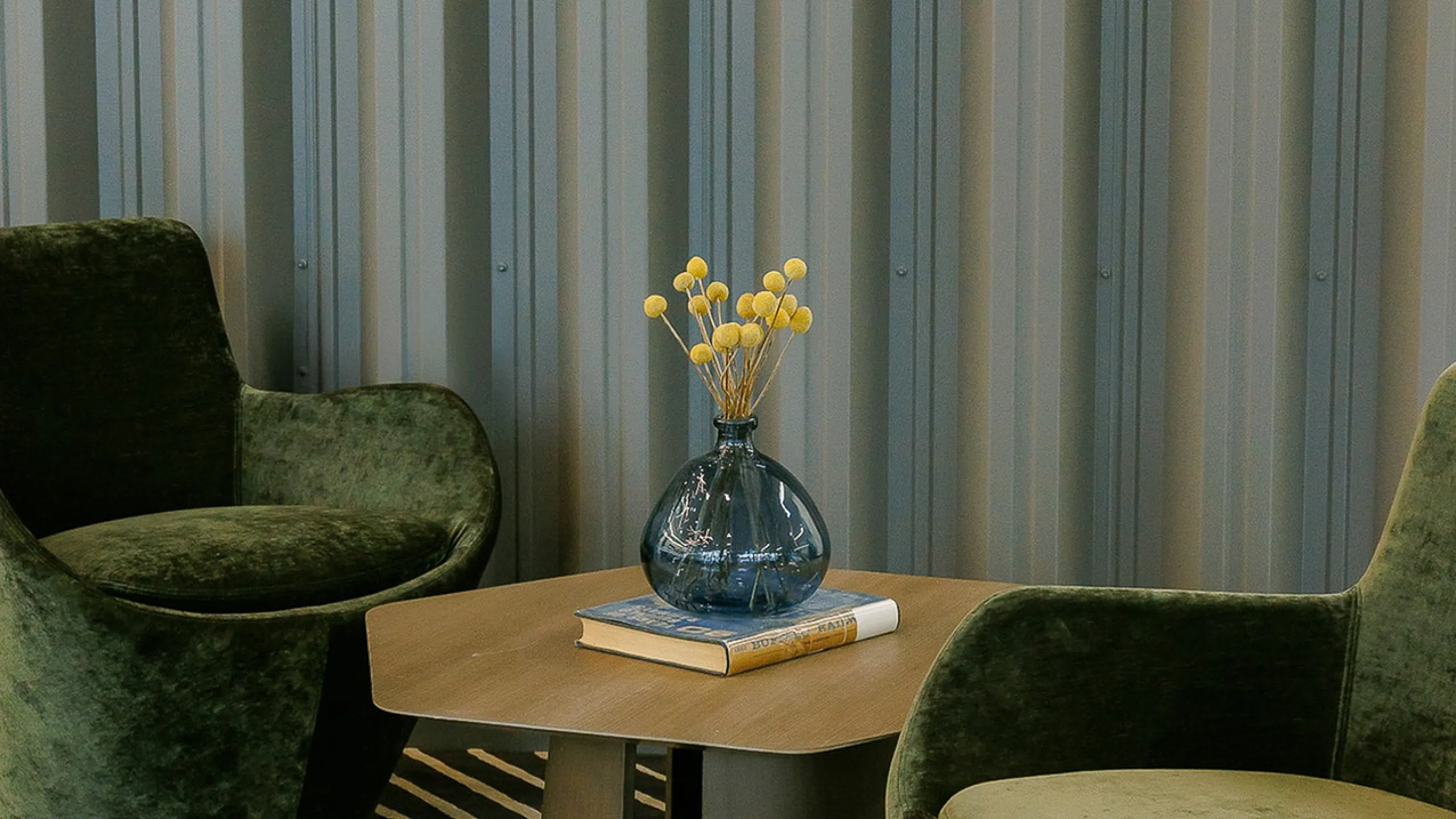 Chic sitting area featuring plush green velvet chairs, a stylish wooden coffee table, and vibrant floral decor set against a modern striped backdrop.