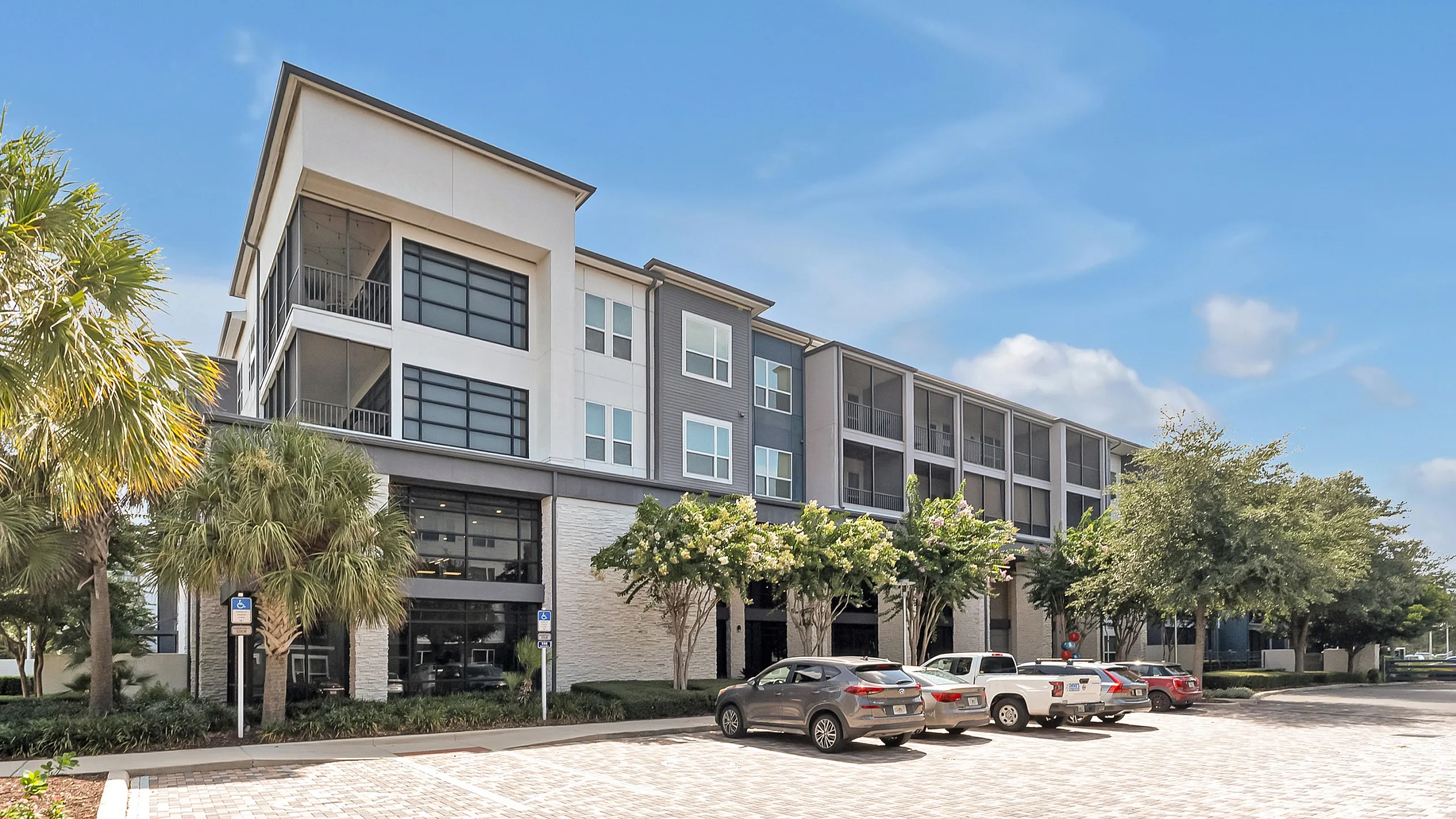 Contemporary apartment complex with sleek architecture, surrounded by lush greenery and ample parking, set against a vibrant blue sky.