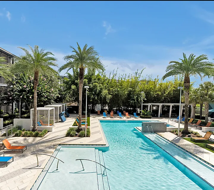 Luxurious apartment complex with a sparkling blue pool, surrounded by palm trees and stylish lounge chairs under a bright sky.
