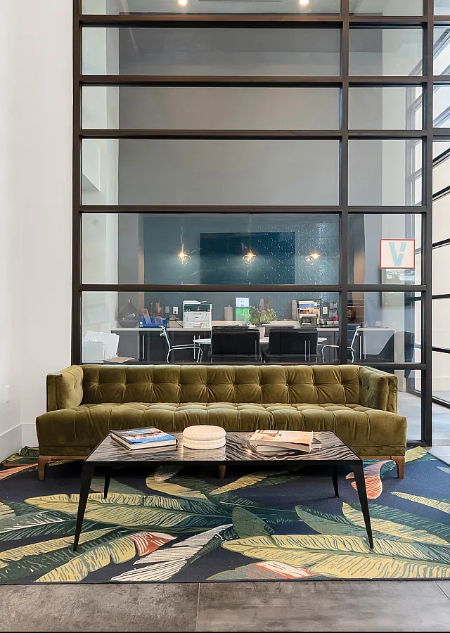 Sophisticated lounge area in modern apartment complex, featuring plush blue and green seating, a stylish rug with leaf patterns, and floor-to-ceiling windows for ample natural light.