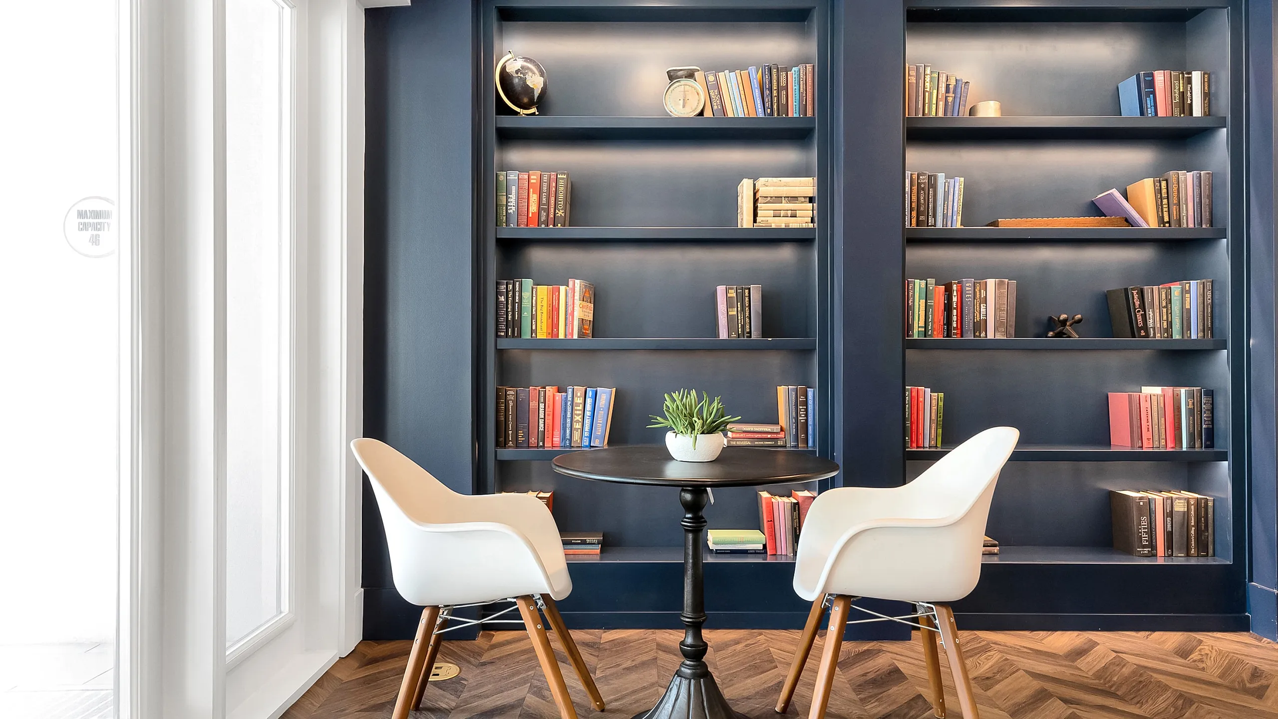 Elegant reading nook with modern white chairs and a sleek round table, framed by a sophisticated built-in bookshelf filled with colorful books.