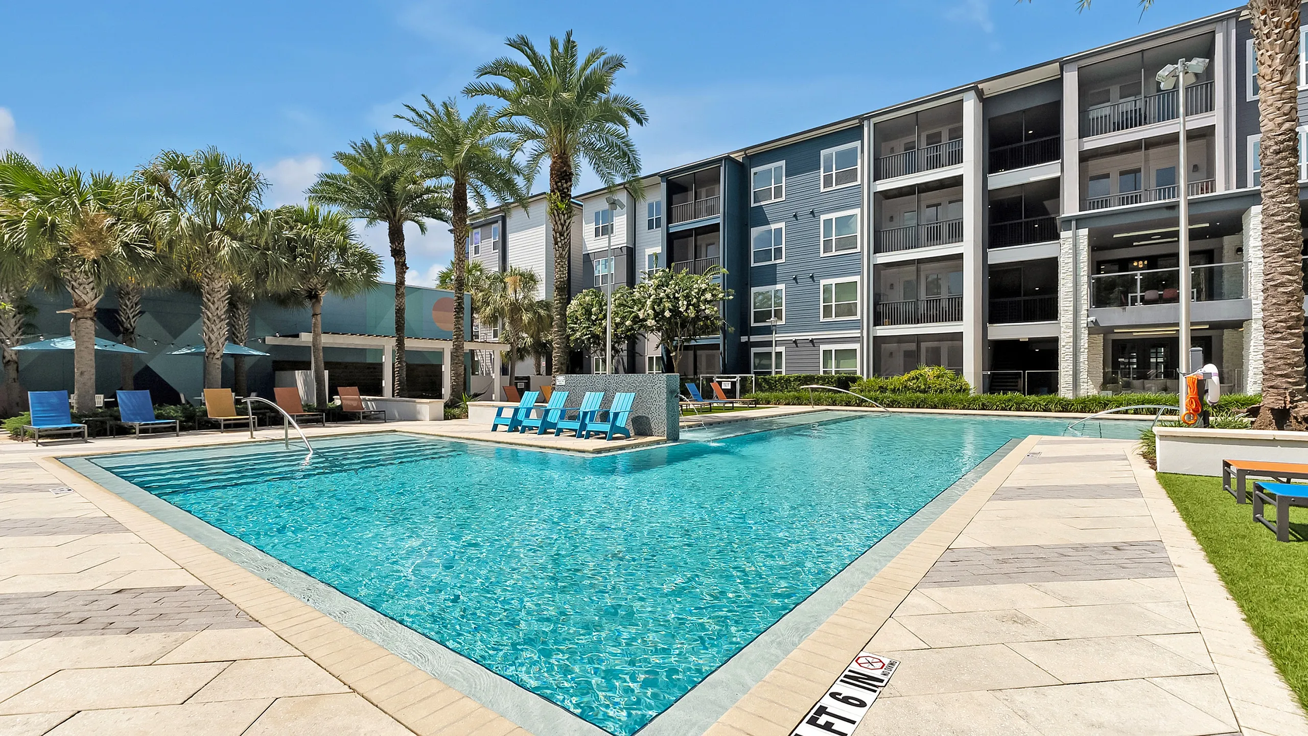 Luxurious apartment complex with a sparkling blue swimming pool, surrounded by palm trees and sun loungers, offering a resort-style living experience.