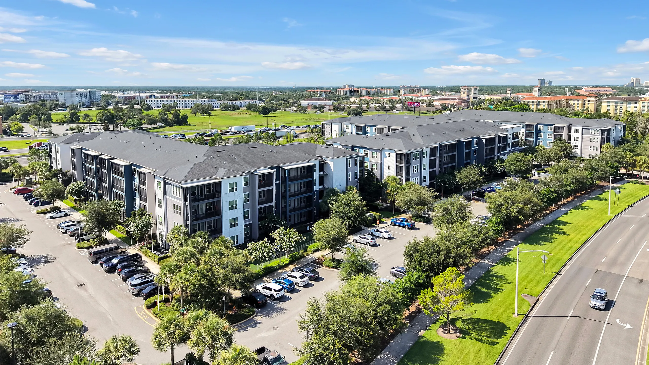 Aerial view of a modern apartment complex surrounded by lush greenery and ample parking, set against a vibrant urban backdrop.