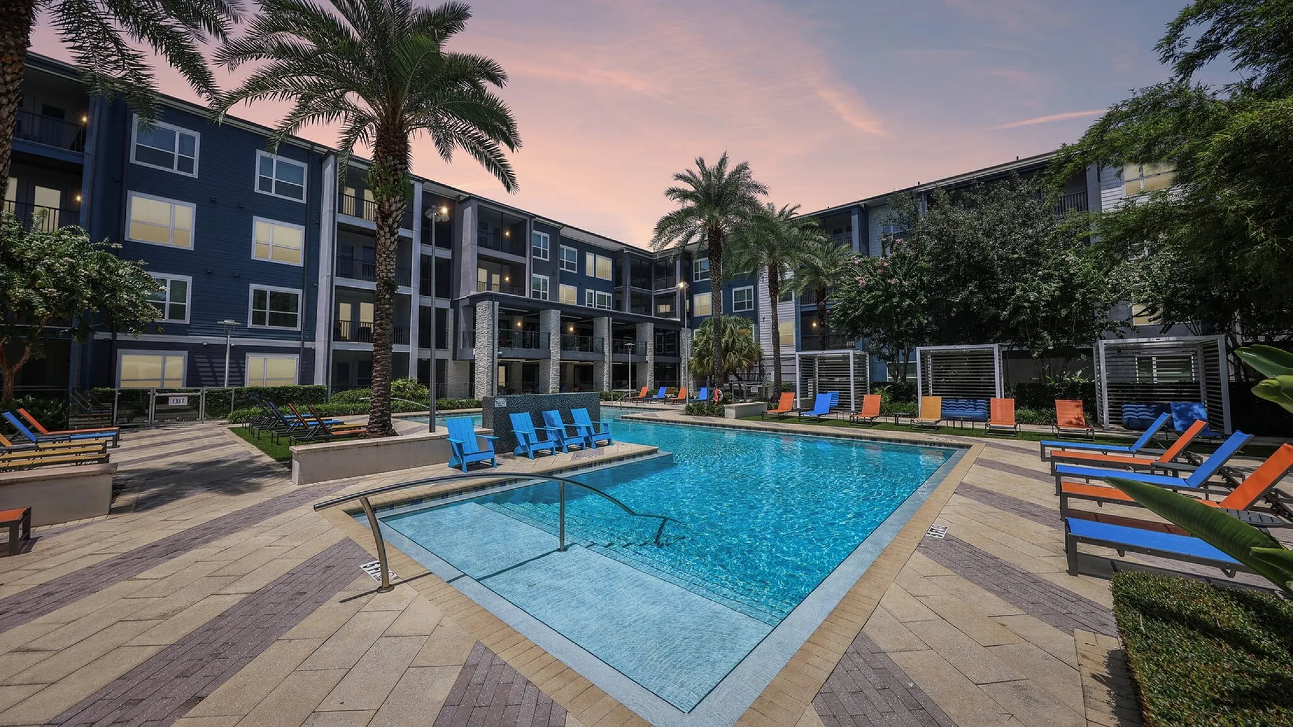 Luxurious apartment complex with a sparkling swimming pool, vibrant lounge chairs, and tall palm trees, perfect for relaxing at sunset.