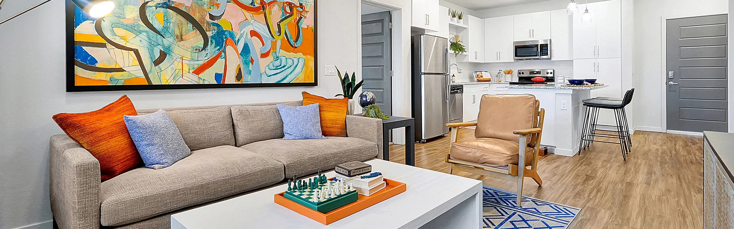 Modern open-concept living room with stylish decor, featuring a contemporary art piece, cozy beige sofa with vibrant pillows, and a sleek white coffee table in front of a chic kitchen area.