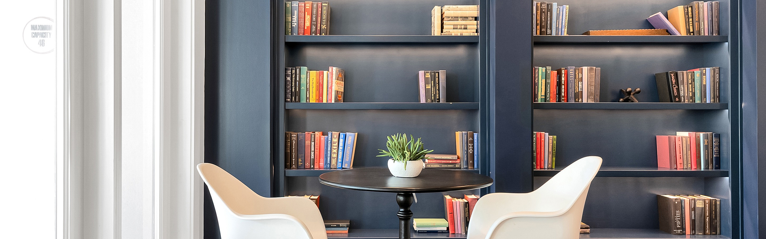 Contemporary reading nook with chic white chairs, a sleek black table, and elegant navy blue bookshelves filled with colorful books.
