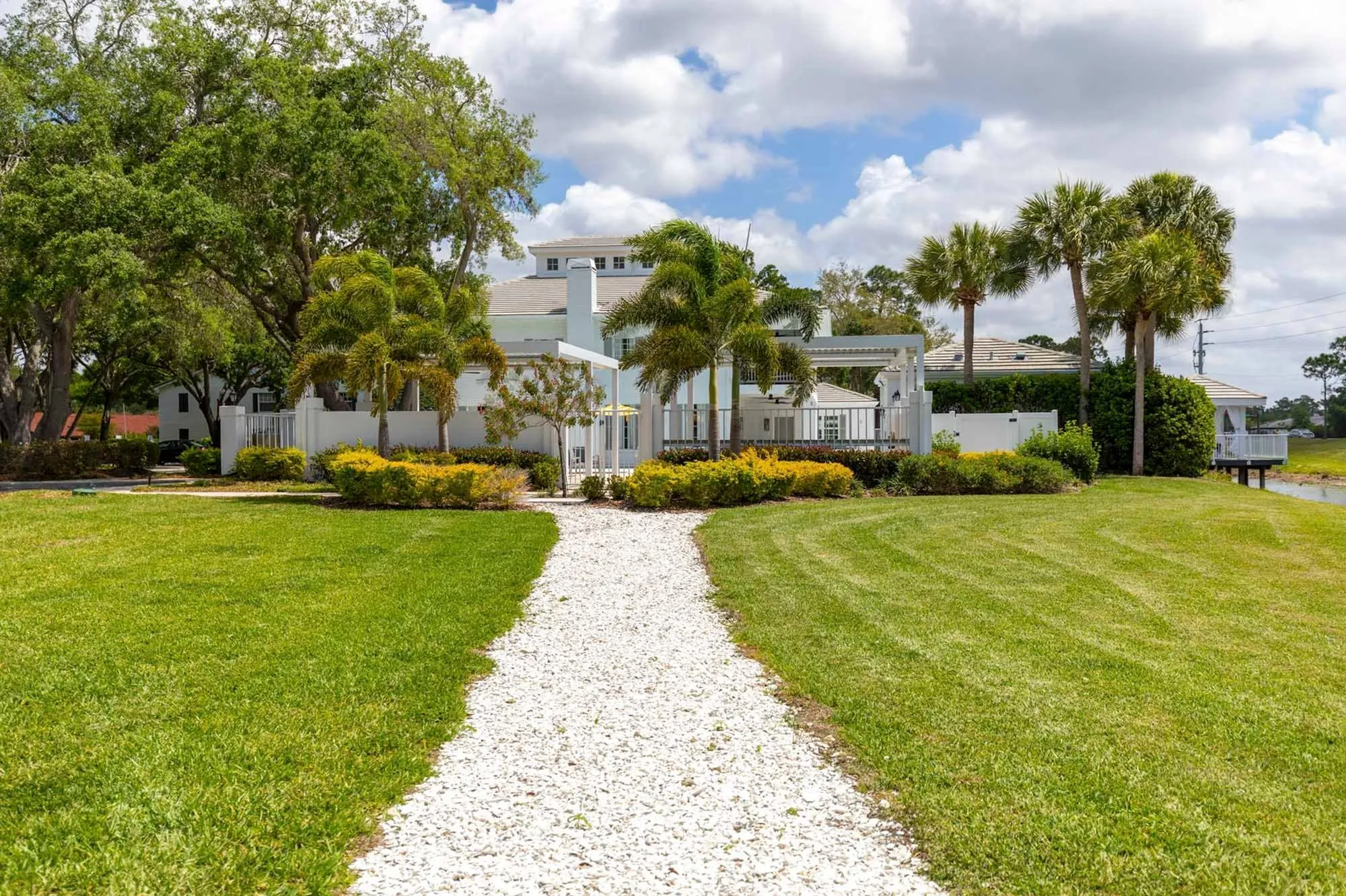 Luxury Apartments for Rent in Sarasota FL - Tradition at Palm Aire - Building Exterior with White Walls, Fence, Grassy Lawn, Palm Trees, and Gravel Path