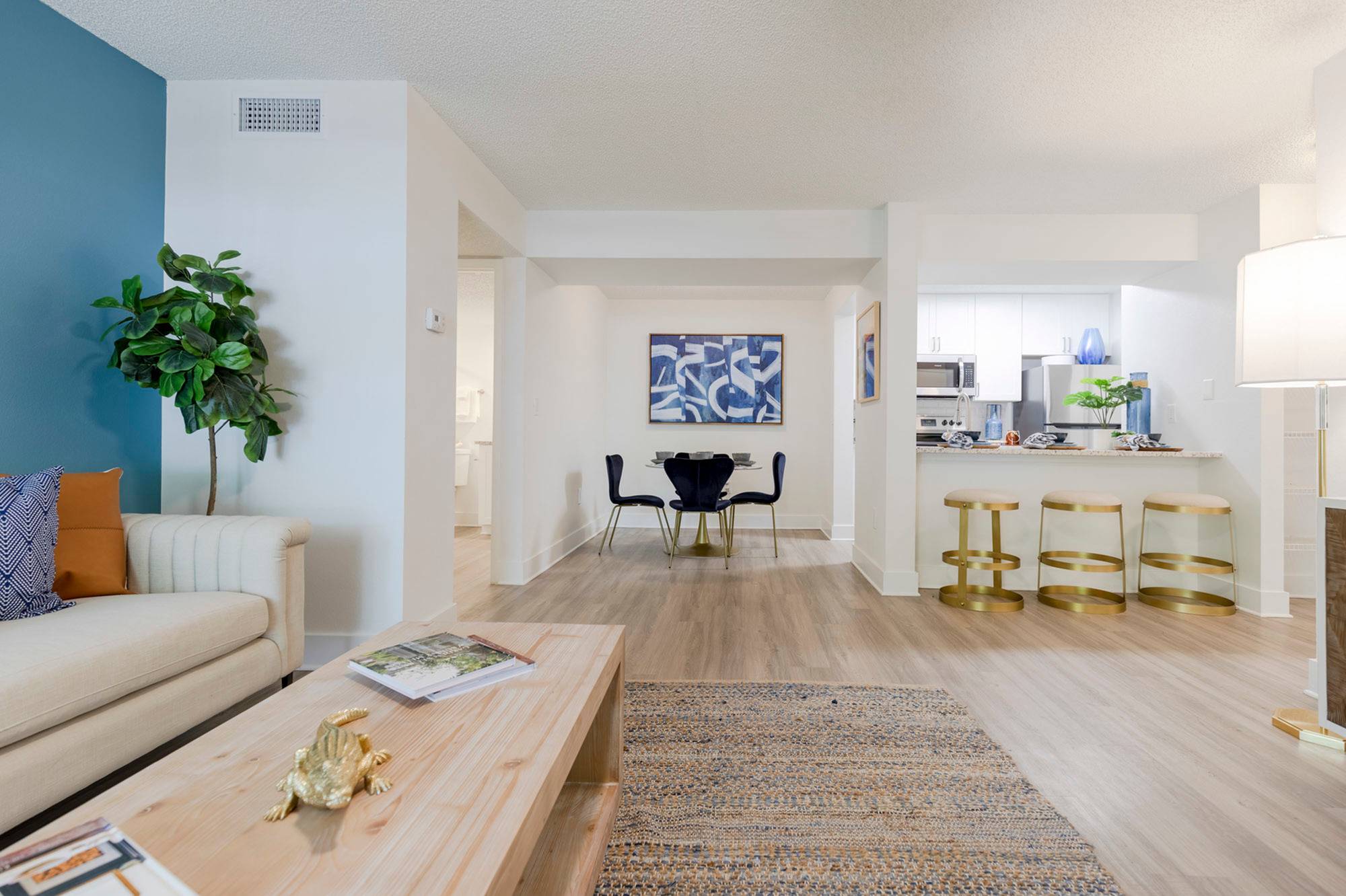 Apartments for Rent in Sarasota, FL - Tradition at Palm Aire - Furnished Living Area with Vinyl Plank Flooring, Area Rug, Sofa, Wooden Cofee Table, Blue Accent Wall, House Plant, and View of Dining Area and Kitchen