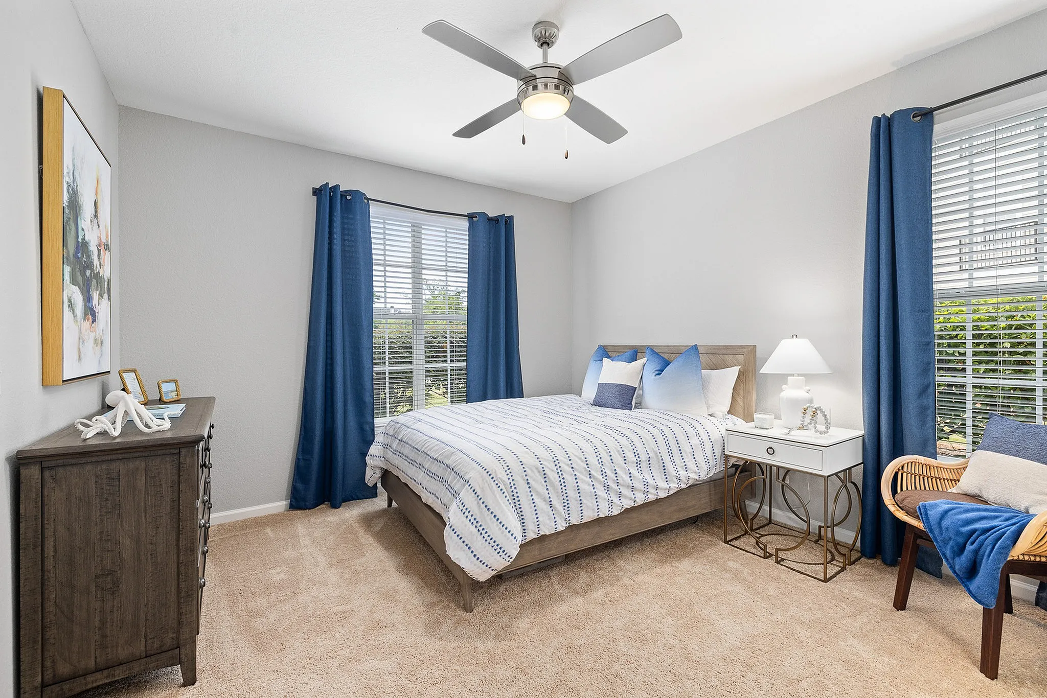 Port Royal, SC, Apartments Near Walmart Supercenter - Preserve at Port Royal - Cozy Bedroom With Ceiling Fan, Blue Curtains, Wood Bed Frame, Nightstand Lamp, And Plush Carpet.