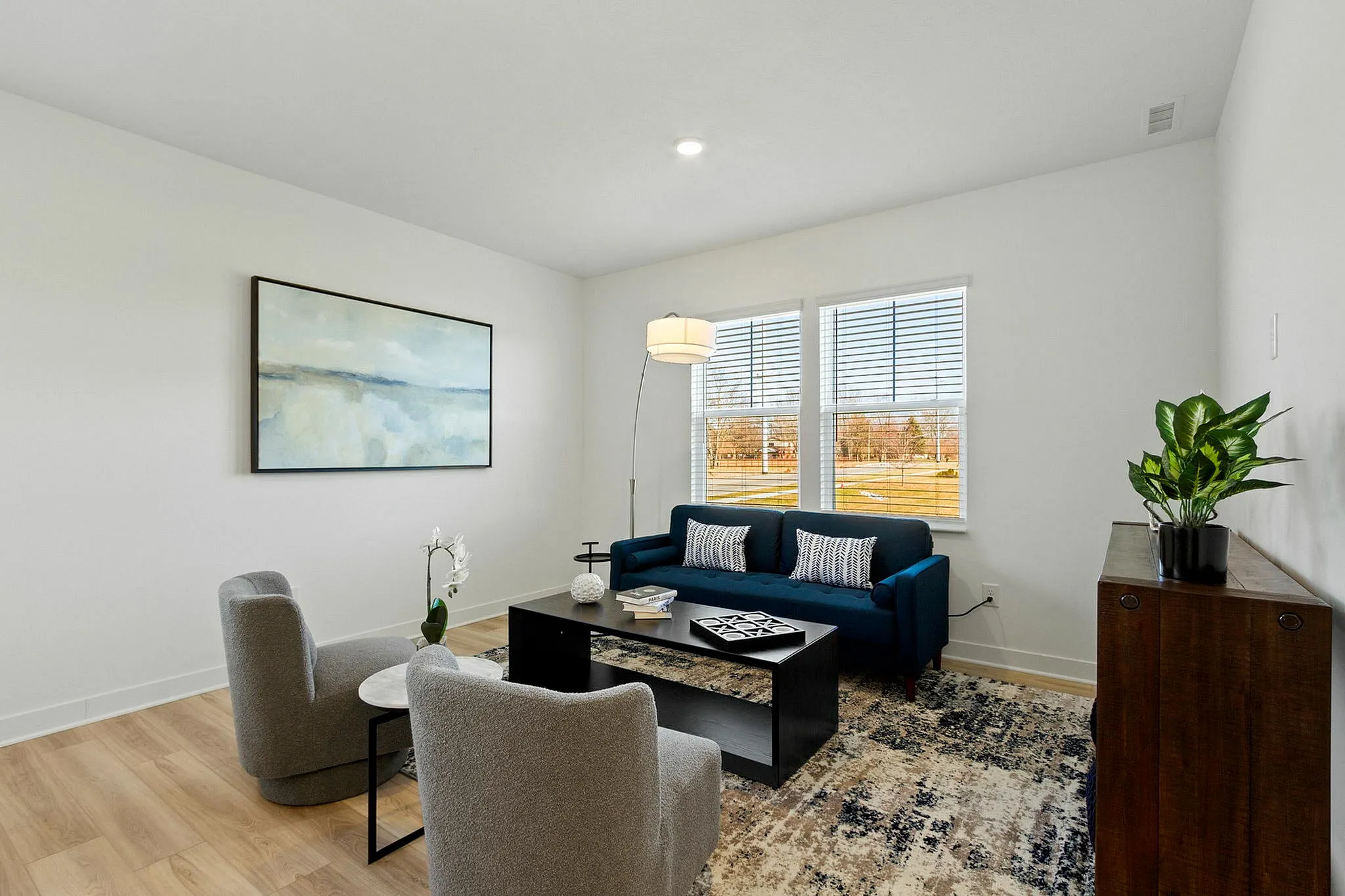 Townhomes with Attached Garage in Brownsburg, IN - Wynne Farms - Living Room With A Blue Sofa, Large Windows, Wood Style Flooring, And A Potted Plant.