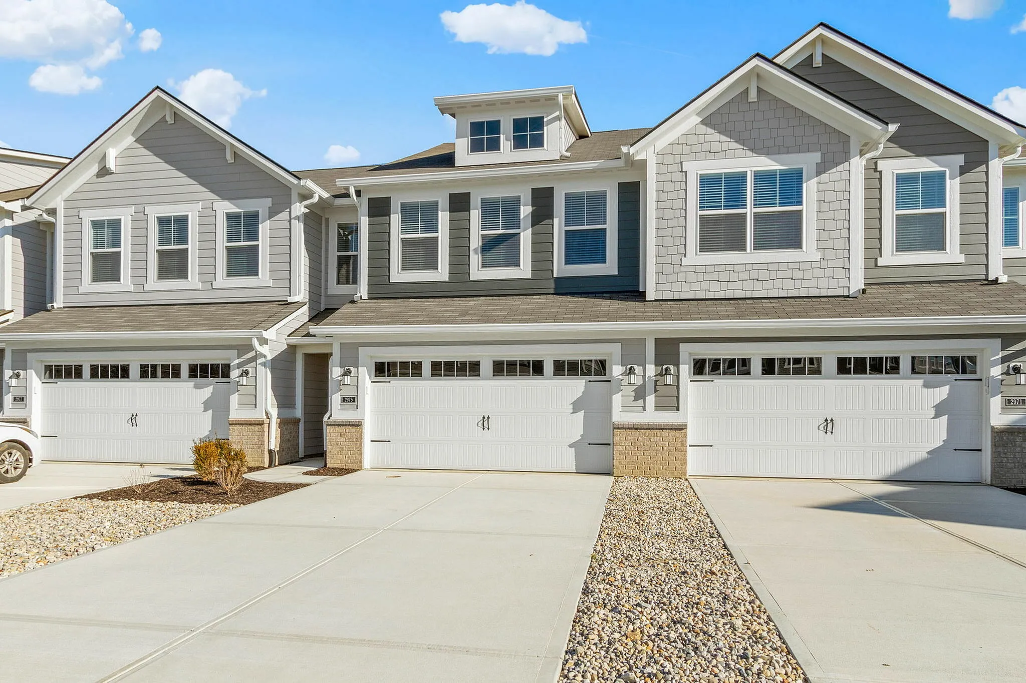 Brownsburg, IN, Townhomes - Wynne Farms - Garage With Gray Siding, White Garage Doors, Large Windows, Private Driveways, And A Bright Blue Sky.